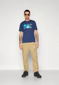 Tumesinist värvi t-särk, millel on värvikas mägimotiiv, kombineeritud beežide vabaajapükstega ja mustade matkasaabastega. Päikeseprillid lisavad sportlikku detaili.