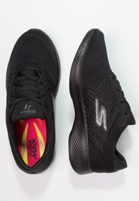 Zwarte sportieve schoenen met een gebreide bovenkant, gestructureerde accenten, dikke rubberen zolen en een levendige roze binnenzool met een zonnestraalpatroon.