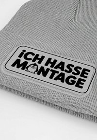Gray ribbed knit beanie featuring a rectangular patch with black text "iCH HASSE MONTAGE" and a small cartoon character.