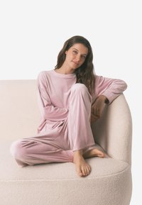 Lichtroze lounge-outfit gemaakt van zacht materiaal, met een lange mouw top en losse broek met ribbelstructuur, zittend op een crème kleurige bank.