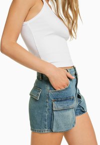 Nainen, joka on pukeutunut valkoiseen ilman hihoja olevaan toppiin ja korkeavyötäröisiin sinisiin farkkushortseihin, joissa on suuret etu- ja sivutaskut. Käsi on sullottu etutaskuun.