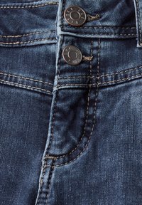 Jeans en denim bleu foncé avec deux boutons en métal à la taille, des coutures jaunes contrastantes et une texture lisse.