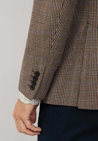 Blazer intrecciato marrone con un motivo a quadri con accenti blu e rossi; quattro bottoni neri sui polsini; camicia bianca e blu a scacchi sotto.