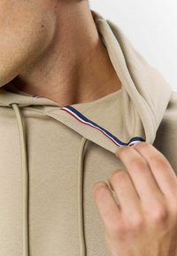 Beige kapucnis pulóver, húzózsinórral, belső részén színes piros és kék csíkkal. Simább textúrájú, laza stílusú.