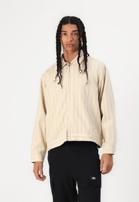 Beige striped jacket with a zip closure, soft texture, and a collared neck. Features side pockets and a relaxed fit. Paired with black pants.