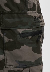 Tissu camouflage avec une poche zippée et une poche à rabat, avec du texte brodé noir sur le rabat supérieur.