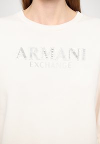Felpa bianca con la scritta "ARMANI EXCHANGE" in argento e borchie decorative. Tessuto liscio e design a girocollo.