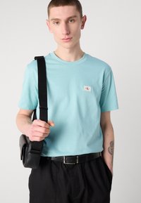 Világoskék pamut póló rövid ujjakkal, kerek nyakkivágással és kis logóval, fekete nadrággal és fekete crossbody táskával párosítva.