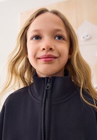 Veste noire zippée avec un col montant, fabriquée en tissu doux. Le design présente une texture subtile et une fermeture éclair centrale.