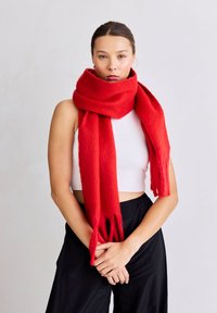 Rode wollen sjaal met een zachte textuur, lange lengte en franjes aan de uiteinden, losjes gedragen. Gecombineerd met een wit shirt en zwarte broek.