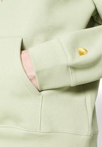 Felpa verde chiaro con tasca frontale, polsini a coste e un piccolo logo ricamato in oro sulla manica. Tessuto morbido e strutturato.