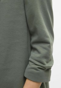 Olijfgroen langgewicht kledingstuk met een zachte textuur, voorzien van opgerolde manchetten en een comfortabele pasvorm. Zomen zijn zichtbaar langs de zijkant.