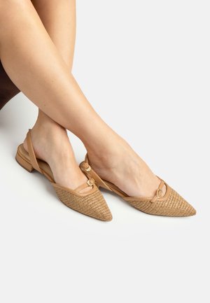 Piede destro con scarpa piatta intrecciata color cuoio con punta appuntita e cinturino a slingback con fibbia, piede sinistro a piedi nudi, incrociati su sfondo bianco.