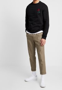 Felpa nera con ricamo di rosa rossa, pantaloni beige e sneakers bianche. Camicia bianca indossata sotto la felpa. Texture liscia.