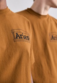 Brązowe t-shirty z bawełny mają czarny graficzny logo z napisem "Aries". Materiał wydaje się miękki, z klasycznym okrągłym dekoltem.