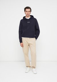 Námornícky mikina s kapucňou a šnúrkou, s predným vreckom, s bielym logom "GUESS". Skombinovaná s béžovými nohavicami a bielymi teniskami, postavená na bielom pozadí.