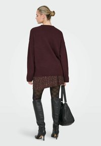 Przesadny bordowy sweter noszony na wzorzystej sukience, czarnych rajstopach i wysokich czarnych skórzanych botkach; trzyma czarną torbę.