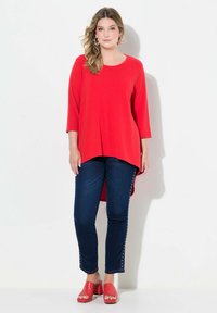 Top rosso a maniche lunghe con scollatura rotonda e orlo asimmetrico. Abbinato a jeans skinny blu scuro con dettagli in borchie sui lati. Sandali con tacco wedge rossi.