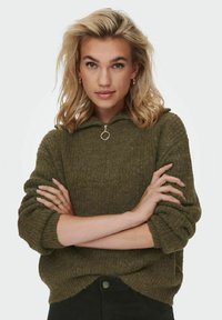 Pull en maille vert olive avec un col haut et une fermeture éclair, présentant un motif texturé et une coupe décontractée.