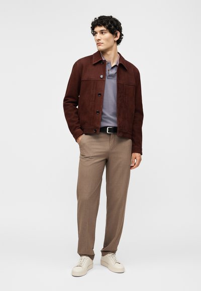 Άνδρας που φορά καφέ κουμπωτό μπουφάν, γκρι πόλο πουκάμισο, μπεζ παντελόνι με μαύρη ζώνη και λευκά αθλητικά παπούτσια, στέκεται μπροστά σε απλό φόντο.