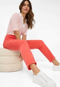 Rot gestreiftes kurzärmliges Poloshirt mit einem weißen Kragen, kombiniert mit hochgekrempelten leuchtend korallenfarbenen Hosen und weißen Sneakers auf einem strukturierten beigefarbenen Hocker.