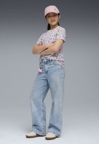 Ljuslila t-shirt med abstrakt mönster, ljusblå baggy jeans, rosa keps och vita sneakers med bruna sulor och rosa pom-pom-accessoar.