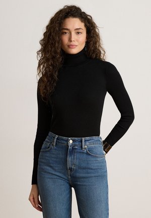 Giovane donna con lunghi capelli ricci che indossa un maglione nero a collo alto e jeans blu a vita alta, in piedi davanti a uno sfondo semplice.