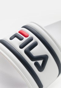 Infradito bianche con dettagli neri e un logo FILA in gomma ben visibile; presenta strisce orizzontali decorative in nero e rosso.