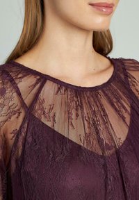 Top in pizzo prugna con scollatura trasparente e motivi floreali, tessuto texturizzato e fodera interna liscia. Effetto drappeggiato elegante.
