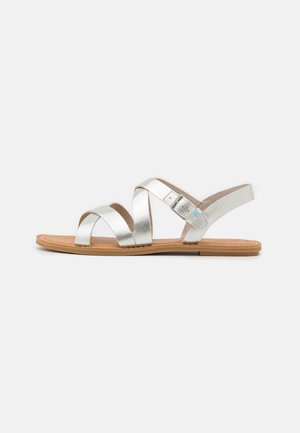 Zilverkleurige metallic sandalen met platte zool, voorzien van kruisbandjes en een verstelbare enkelriem op een beige voetbed.