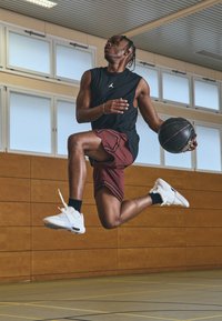 Una persona salta en un gimnasio, vistiendo una camiseta sin mangas negra, pantalones cortos burdeos con patrones y zapatillas de baloncesto blancas, sosteniendo un balón de baloncesto negro.
