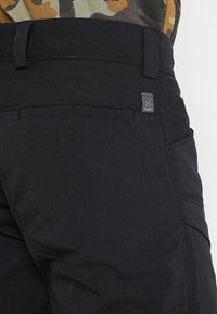 Gros plan sur un pantalon noir avec passants de ceinture, une poche passepoilée à l'arrière et une petite étiquette en tissu près de la taille.