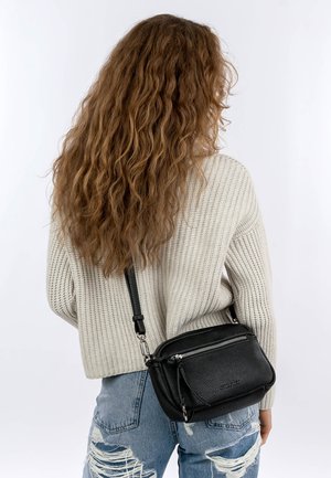 Persona con cabello largo y rizado que lleva un suéter crema de canalé, jeans azules rotos y un bolso cruzado de cuero negro con cremalleras plateadas.