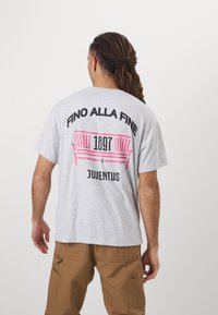 Šedé bavlnené tričko s čiernymi a ružovými grafikami na chrbte, obsahujúce nápisy "Fino Alla Fine," "1897" a "Juventus." Krátke rukávy a voľný strih.