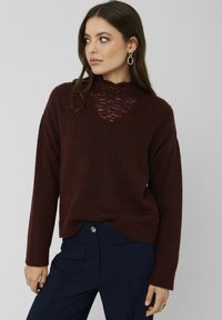 Pull en tricot bordeaux avec un décolleté en V et des détails en dentelle au niveau de la poitrine ; coupe ample avec des poignets et un ourlet côtelés ; porté sur un pantalon bleu marine.