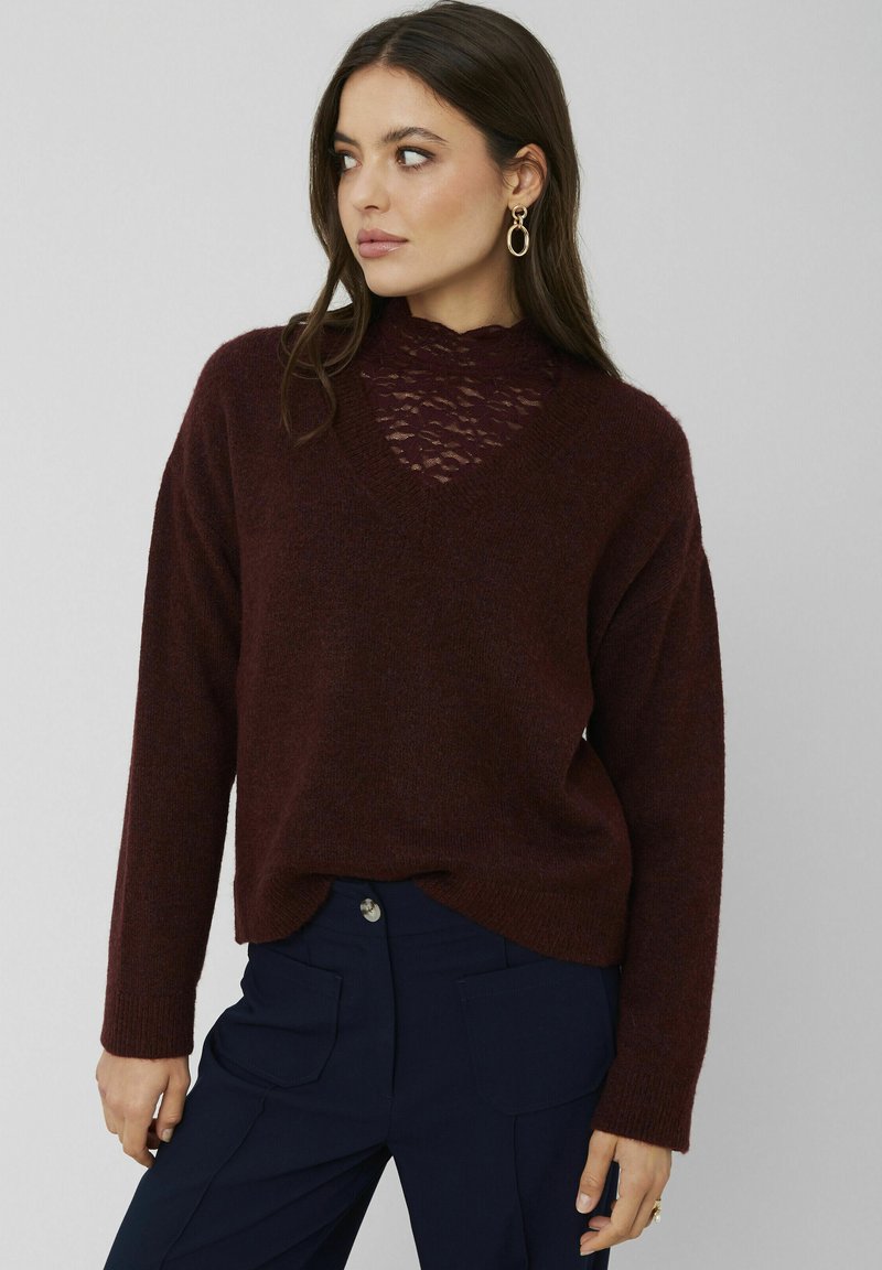 Pull en tricot bordeaux avec un décolleté en V et des détails en dentelle au niveau de la poitrine ; coupe ample avec des poignets et un ourlet côtelés ; porté sur un pantalon bleu marine.