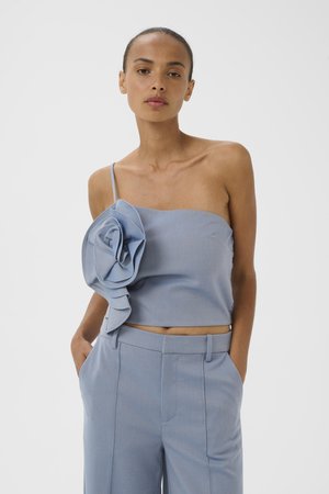 Femme portant un crop top bleu clair une épaule avec une grande fleur en tissu et un pantalon taille haute assorti, les mains dans les poches.