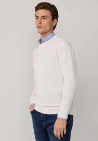 Witte gebreide trui met een V-hals, geribbelde manchetten en zoom. Gelaagd over een blauw-wit gestreept overhemd met kraag. Draag het met donkerblauwe jeans.