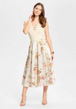 Kvinde iført en cremefarvet blomster-midi kjole med omslags-style bodice, matchende bælte og gyldne stropper høje hæle, poserer med hånd nær ansigtet.
