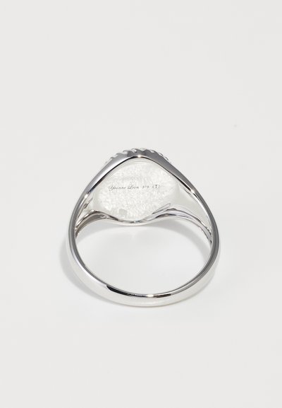 Anello in argento sterling con una superficie liscia e lucida, caratterizzato da un bordo testurizzato e un ampio centro bianco a forma ovale. Dettagli incisi visibili.