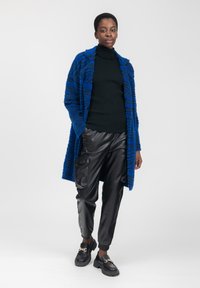 Donna che indossa un cappotto blu a fantasia, un dolcevita nero con ruches, pantaloni cargo in pelle nera e mocassini neri con catene dorate.