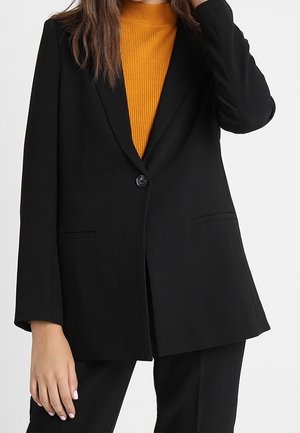Vrouw draagt een zwarte blazer over een mosterdkleurige geribbelde coltrui, met bijpassende zwarte broek, hand bij het gezicht en een ontspannen houding.