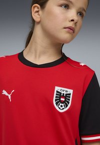 Κόκκινη φανέλα ποδοσφαίρου με μαύρες λεπτομέρειες, που διαθέτει στρογγυλή λαιμόκοψη, κεντημένο λογότυπο Puma και το έμβλημα της ÖFB στο κέντρο. Υφασμα με υφή.