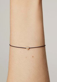 Pulsera de cordón negra con un pequeño charm circular dorado adornado con cristales transparentes, llevada en una muñeca de tono de piel claro.