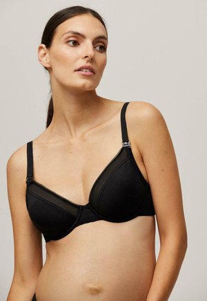 Sujetador negro que presenta una mezcla de tejido suave y paneles de malla transparente, con una forma de copa triangular y correas ajustables, acentuado por un ribete decorativo.