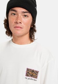 Camiseta blanca de manga corta con un colorido logotipo gráfico en el pecho. El modelo lleva un gorro de punto negro. Tejido texturizado.
