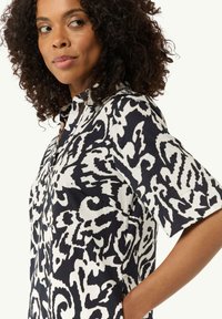 Kurzärmeliges Hemd aus schwarz-weißem Stoff mit abstrakten, wirbelnden Mustern, Knopfverschluss vorne und Seitentaschen.