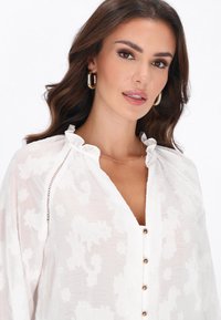 Blusa blanca con estampado floral, cuello con volantes, detalle de botones y textura sutilmente transparente. Acentuada con herrajes dorados brillantes.
