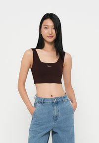 Brun cropped topp med brede stropper og firkantet hals, med en liten hvit logo på forsiden. Kombinert med lyseblå denimbukser.