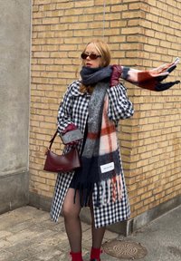 Manteau à carreaux bleu et blanc, écharpe grise avec des accents orange et bleu marine, sac à main bordeaux, et chaussettes rouges. Cheveux bruns, lunettes de soleil et gants.